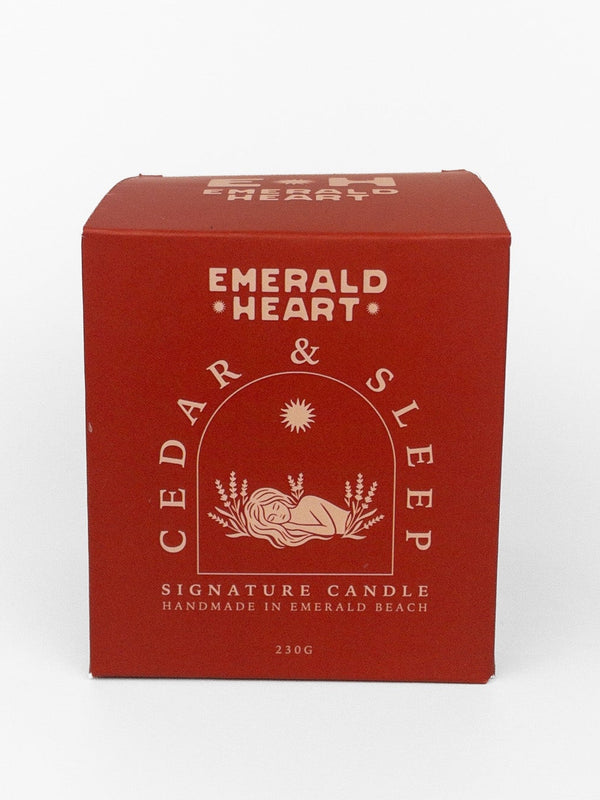 A red rectangular box for Emerald Heart Cedar & Sleep Signature Candle, featuring cream-colored text and a sun-and-book illustration, photographed on a clean white background.